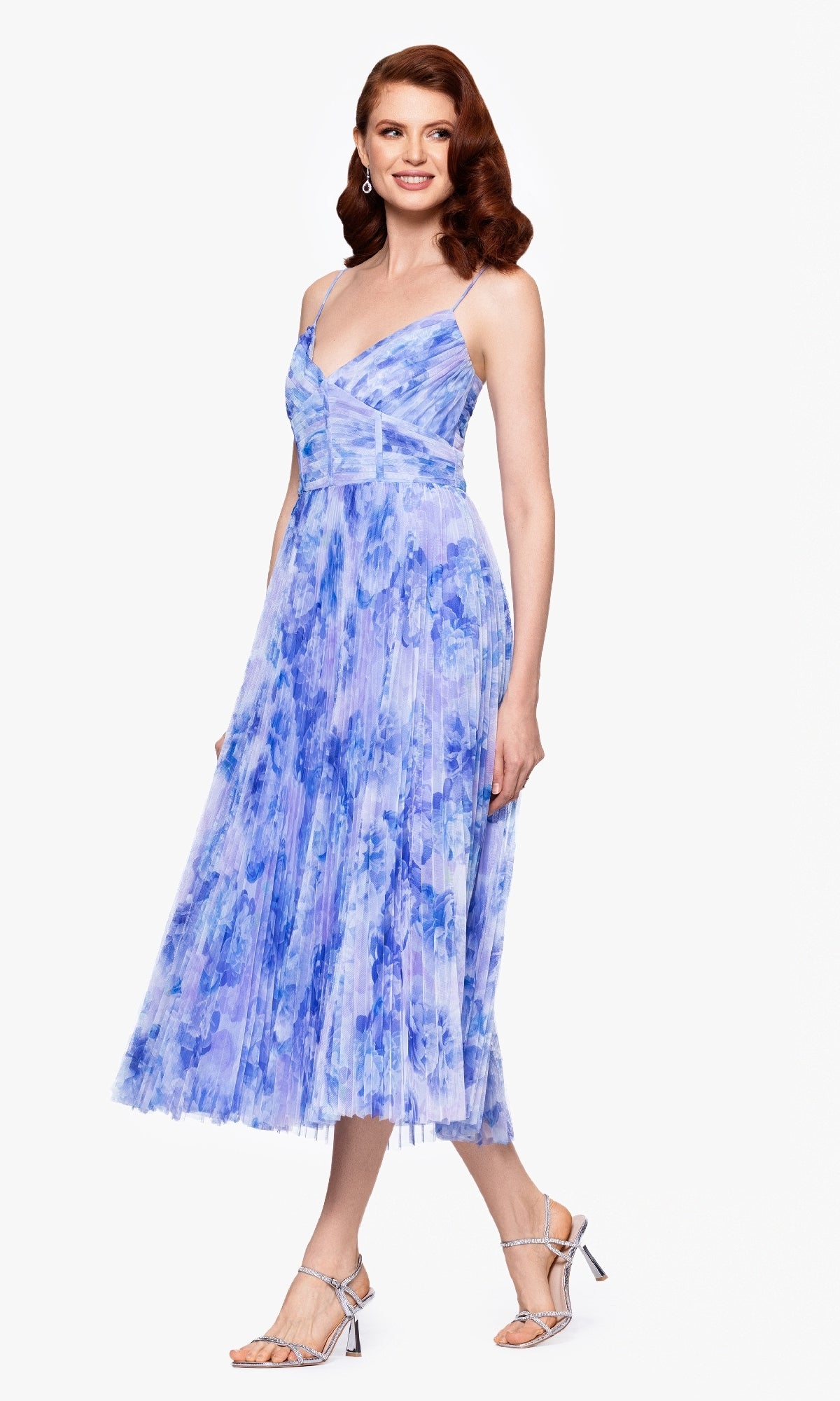 A woman wears the Betsy & Adam A27053 Midi Wedding Guest Dress with a pleated a-line silhouette, light blue and purple floral print, spaghetti straps, and a structured bodice, paired with silver high-heeled sandals against a white background.